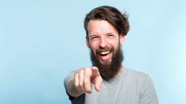 ひげを生やした男性が笑いながらカメラのレンズを指さしています